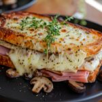 A Golden Brown Croque Monsieur Sandwich With Melted Raclette Cheese, Ham, Herbs, And Mushrooms, Served On A Black Plate.