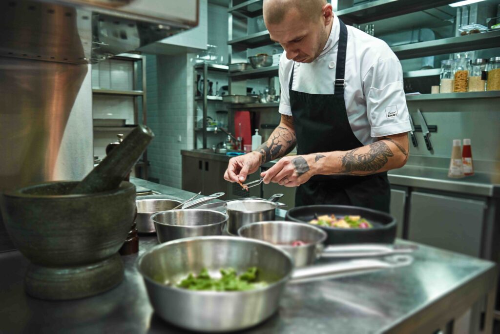 Un chef professionnel concentré dresse soigneusement une assiette dans une cuisine moderne, entouré d’ustensiles et d’ingrédients, illustrant la précision et la créativité rendues possibles grâce aux sauces prêtes à l’emploi Cuisin’Émoi.