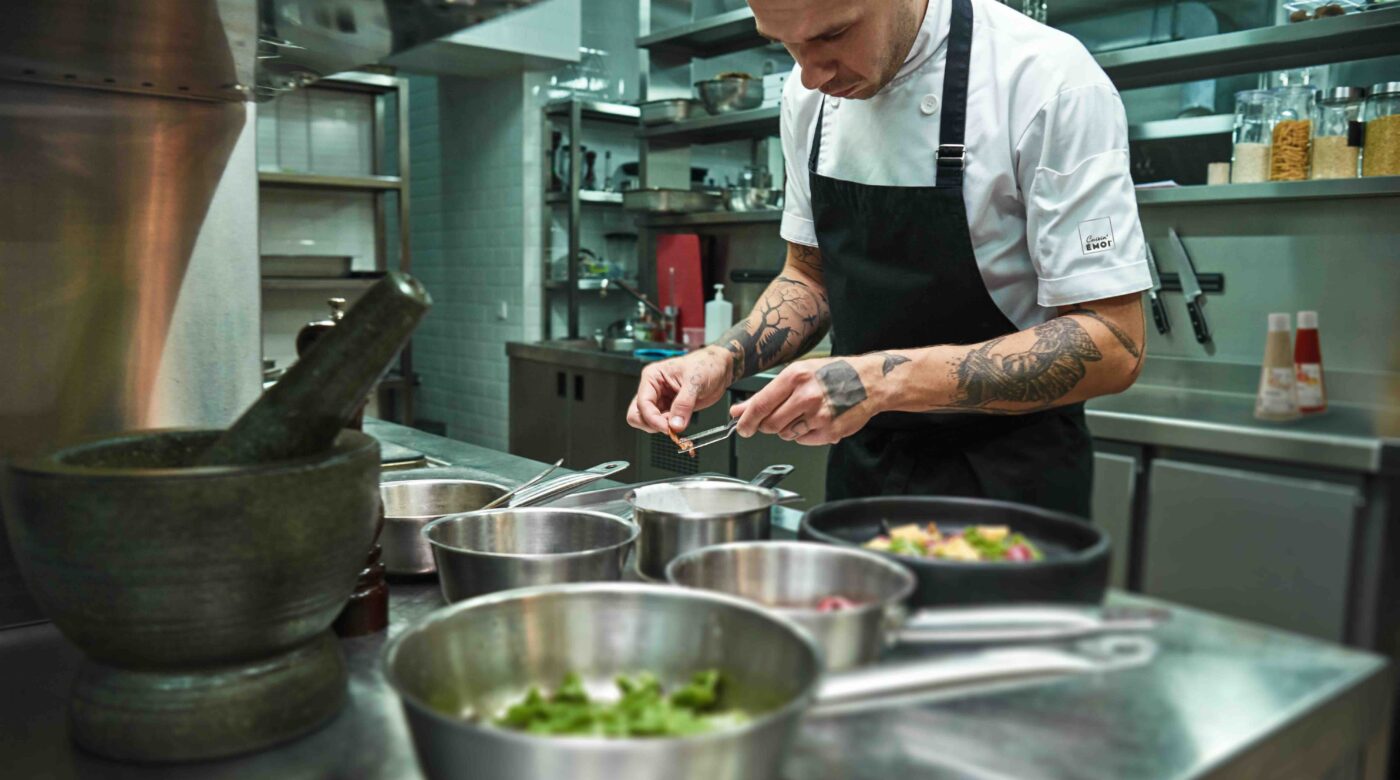 Un chef professionnel concentré dresse soigneusement une assiette dans une cuisine moderne, entouré d’ustensiles et d’ingrédients, illustrant la précision et la créativité rendues possibles grâce aux sauces prêtes à l’emploi Cuisin’Émoi.