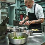 Un chef professionnel concentré dresse soigneusement une assiette dans une cuisine moderne, entouré d’ustensiles et d’ingrédients, illustrant la précision et la créativité rendues possibles grâce aux sauces prêtes à l’emploi Cuisin’Émoi.