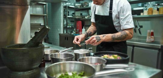 Un chef professionnel concentré dresse soigneusement une assiette dans une cuisine moderne, entouré d’ustensiles et d’ingrédients, illustrant la précision et la créativité rendues possibles grâce aux sauces prêtes à l’emploi Cuisin’Émoi.