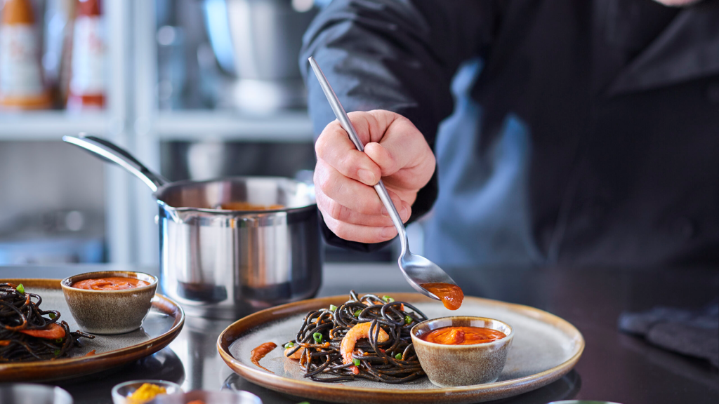 Chef en cuisine dressant un plat et tenant une cuillère de sauce Cuisin'Emoi.
